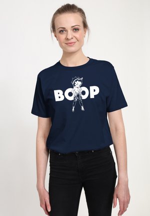 Žena se světlými vlasy, která má na sobě tmavě modré tričko s kreslenou postavičkou a bílým nápisem „BOOP“, doplněné černými kalhotami.