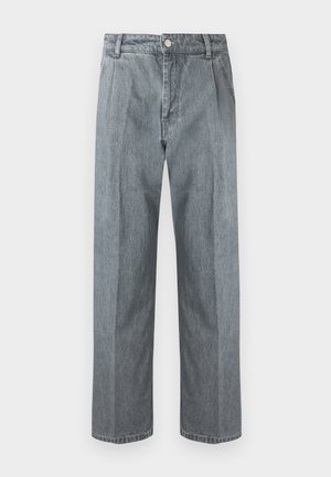 Osoba nosząca czarne okulary przeciwsłoneczne, kremowy sweter z granatowymi paskami, szerokie szare dżinsy, czarne baleriny, trzyma dużą czarną skórzaną torbę na białym tle.