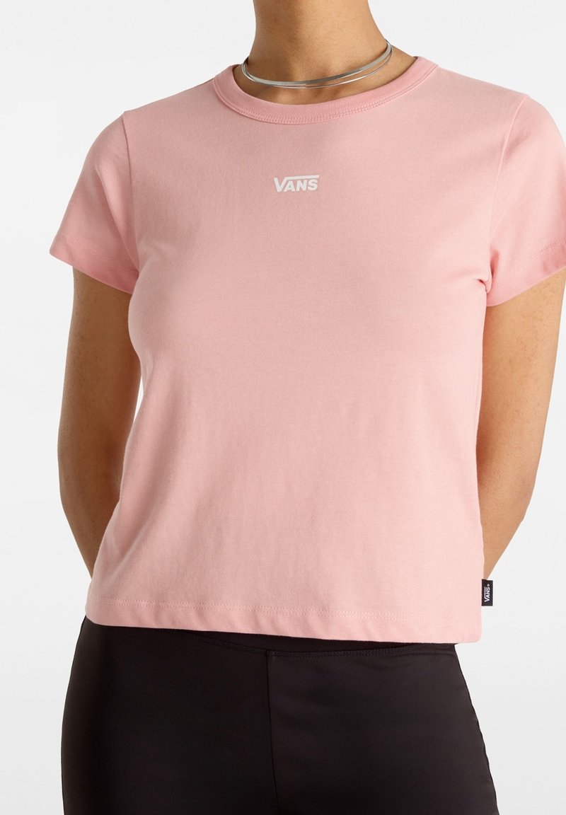 Růžové tričko s krátkým rukávem vyrobené z měkké látky, s bílým logem Vans na hrudi a rovnou spodní částí. Pod ním jsou nošeny černé kalhoty.