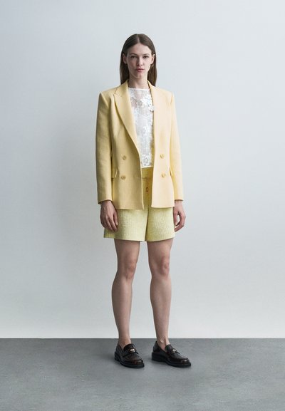 Femme debout contre un mur uni, portant un blazer jaune pâle, un short texturé assorti, un top en dentelle blanche et des mocassins noirs.