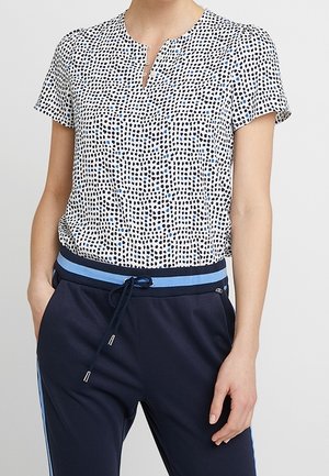 Kobieta ubrana w białą bluzkę z krótkim rękawem z czarnymi i niebieskimi kropkami, w połączeniu z granatowymi spodniami z niebieskim paskiem w paski i sznurkiem do ściągania.