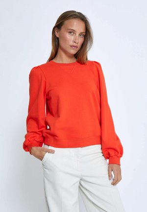 Světlý oranžový svetr s dlouhými, vycpanými rukávy, kulatým výstřihem a mírně zkráceným lemem. V kombinaci s bílými kalhotami.