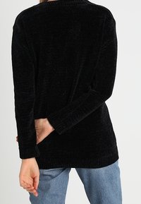 Černý svetr s dlouhým rukávem s texturovaným vzorem, volným střihem a žebrovanými detaily, doplněný světle modrými džínami. Pohled zezadu odhaluje jednoduchý design.