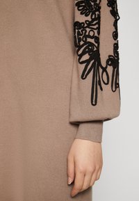 Strikket kjole i beige stof med intrikate sorte broderier på ærmet, der har tekstureret detalje og en løs pasform ved håndleddet.