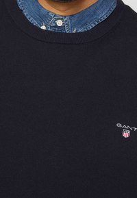 Granatowy sweter robiony na drutach z okrągłym dekoltem, z deninowym kołnierzykiem pod spodem oraz logo GANT na piersi. Gładka faktura i dopasowany fason.