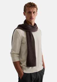 Bruine ribgebreide sjaal van zacht materiaal, gedragen over een lichtbeige trui, met een lange rechthoekige vorm en een gladde textuur.