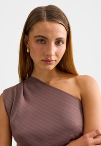 Νέα γυναίκα με ίσια ανοιχτό καστανά μαλλιά, φοράει ένα ρούχο με έναν ώμο σε μωβ απόχρωση με λεπτές λευκές ρίγες και ένα χρυσό σκουλαρίκι, ουδέτερο φόντο.