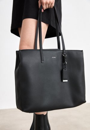 Persoon houdt grote zwarte Calvin Klein tas met dubbele handvatten vast, draagt een zwarte jurk en zwarte enkellaarzen tegen een lichte achtergrond.