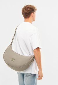 Világosszürke anyagú, kerek formájú sling táska. Erős pánttal és egy kis címkével rendelkezik az elején. Simá szövet és minimalista dizájn jellemzi.