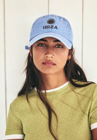 Light blue cotton cap with a sun logo and "IBIZA" text in brown, worn with a green t-shirt featuring white accents.
