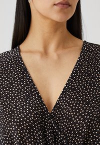 Μαύρη πουκαμίσα με σχέδιο που συνδυάζει υφή υφάσματος με μικρές κρέμες κουκίδες, βαθύ V-ανοιχτό και απαλά drapée στοιχεία στο σχέδιο.