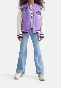 Purple varsity jacket with cream sleeves, striped cuffs, and embroidered patches. Worn over a white shirt and paired with light blue jeans.