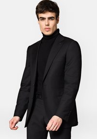 Czarny garnitur o dopasowanym kroju z wyraźnymi klapami, w zestawieniu z czarnym golfem. Gładki materiał z delikatnym połyskiem i eleganckim krojem.