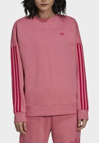 Rosa sweatshirt med långa ärmar, med tre mörkare rosa vertikala ränder på varje arm och en liten logotyp på bröstet. Mjuk textur.