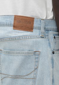 Jeans de mezclilla azul claro con un parche de cuero etiquetado como "Hollister" en la cintura. Presenta costuras naranjas y bolsillos traseros clásicos.