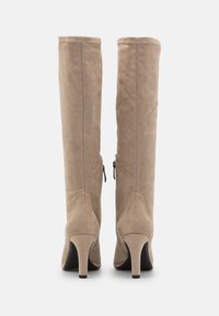 Un paio di stivali alti fino al ginocchio in suede beige con tacchi sottili visti da dietro, mostrando le cuciture e le linguette della cerniera laterale.