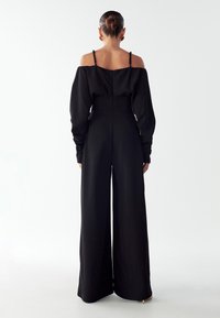 Moteris stovi atsukusi nugarą, aps wearing juodą off-shoulder kombinezoną su ilgomis rankovėmis ir plačiomis kelnėmis prieš paprastą foną.