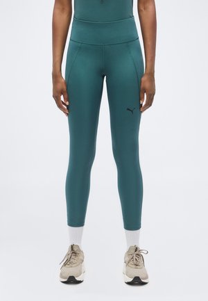 Jambes portant des leggings taille haute bleu canard avec le logo Puma, des chaussettes blanches et des chaussures de course beiges, debout devant un fond uni.