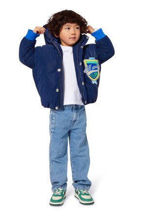 Doudoune bleue avec capuche, boutons-pression et patch. T-shirt blanc, jeans bleu clair, et baskets avec accents verts complètent la tenue.