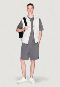 Chaleco acolchado blanco sobre una camiseta gris, combinado con pantalones cortos grises. Calzado: zapatillas blancas. La modelo lleva un bolso negro de hombro.