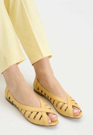 Füße, die blassgelbe, offene, flache Schuhe mit überkreuzten Riemen und Ausschnittdetails tragen, kombiniert mit hellgelben, gekürzten Hosen vor weißem Hintergrund.