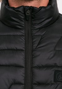 Giacca puffer nera con colletto alto, chiusura a zip e design trapuntato. Logo evidente sull'area del petto sinistro. Tessuto morbido e lucido.