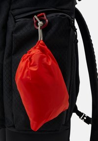 Zaino nero con tessuto a motivo a rombi, dotato di una sacca con coulisse rossa attaccata. Texture liscia con cerniere e spallacci regolabili.