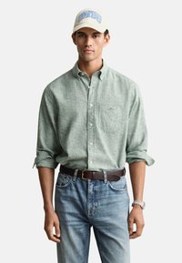 Hellgrünes Hemd mit Knöpfen und hochgekrempelten Ärmeln, blaue Jeans, brauner Gürtel und eine cremefarbene Baseballkappe mit blauem Text. Weißer Hintergrund.