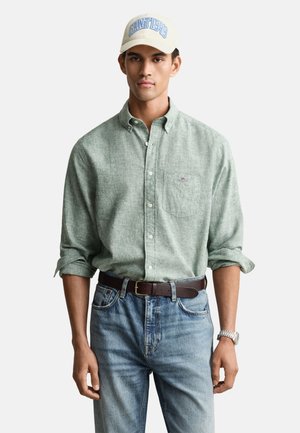 Lysegrøn skjorte med knapper og op rullede ærmer, blå jeans, brunt bælte og en cremefarvet baseballkasket med blå tekst. Hvid baggrund.