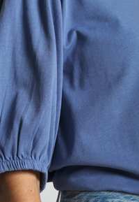 Modrá, strukturovaná blůza s nadýchanými rukávy a elastickými manžetami; hladce se přizpůsobuje tělu, má volný střih a jemný materiál.
