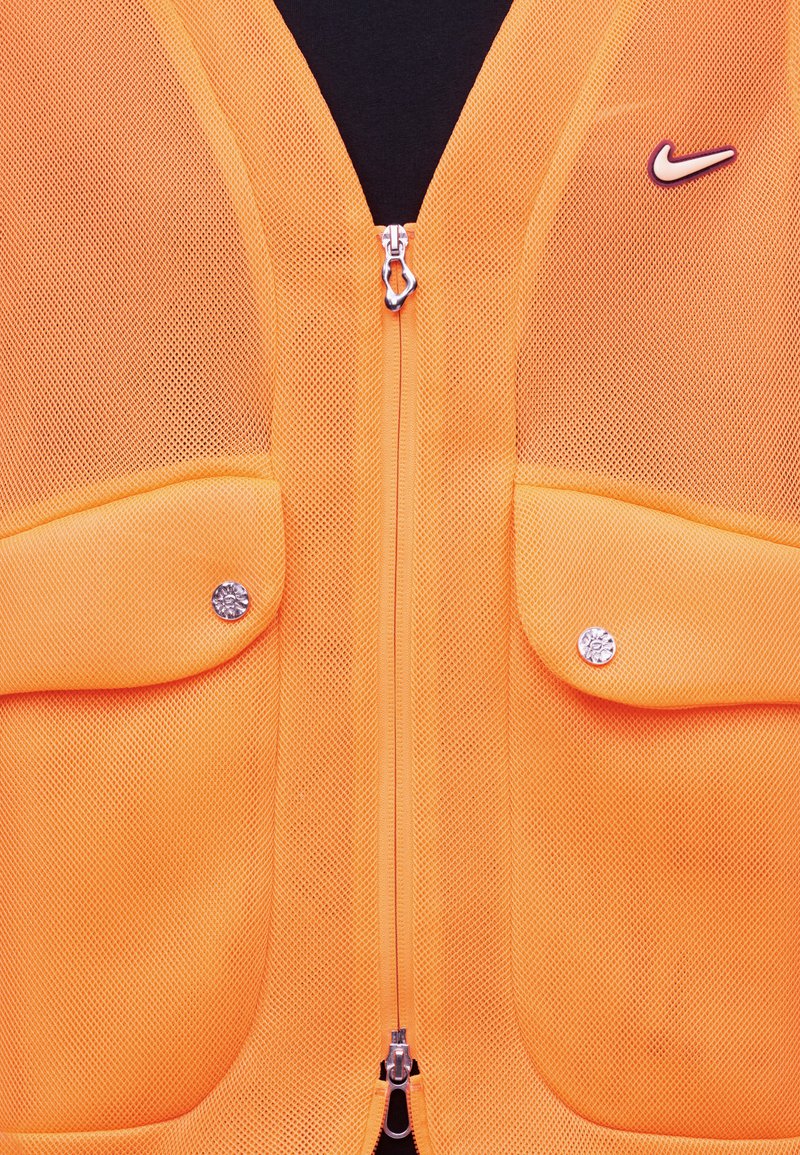 Leuchtend orangefarbenes Netzweste mit Reißverschluss vorne, zwei Klappentaschen mit Metall-Druckknöpfen und einem weißen Nike-Swoosh-Logo auf der Brust.