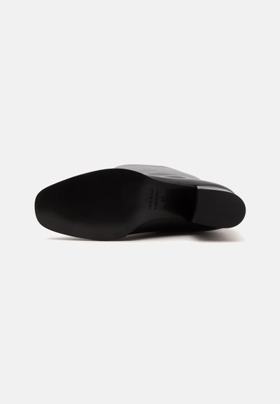 Botte en cuir noir vue par le dessous, mettant en valeur la semelle lisse et le talon carré sur un fond clair uni.
