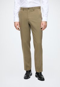 Beige Anzughose mit geradem Schnitt, aus glattem Stoff, mit einem Knopfverschluss vorne und seitlichen Taschen, kombiniert mit schwarzen Schuhen.