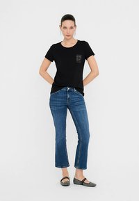 Črna kratka majica s kratkimi rokavi z detajlom prsnega žepa, kombinirana z modrimi zvončastimi jeans hlačami in črnimi balerinkami z teksturiranim vzorcem.