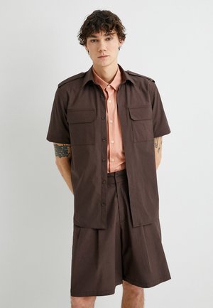 Camisa de botones marrón de manga corta con dos bolsillos en el pecho sobre una camisa de color rosa claro con cuello, combinada con pantalones cortos marrones a juego.
