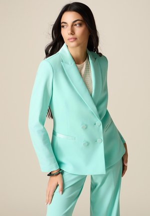 Pitkähiuksinen nainen, joka pukeutunut mintunvihreään kaksiriviseen bleiseriin ja siihen sopiviin housuihin, katsoo sivulle beigeä taustaa vasten.