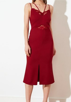 Femme portant une robe midi rouge profond sans manches, avec des détails en dentelle au niveau du décolleté et de la taille, et une fente sur le devant.