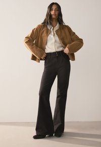 Hnedá semišová bunda so širokými rukávmi v kombinácii s čiernymi zvonovými džínami s detailmi na gombíky a bielou pruhovanou blúzkou.