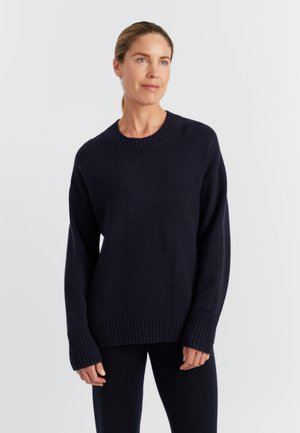 Pull en tricot bleu marine avec un col ras du cou et des poignets côtelés. Texture douce et coupe décontractée, associé à un pantalon sombre assorti.