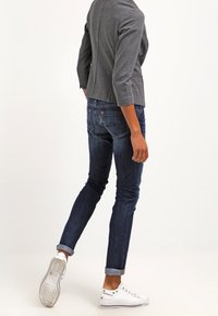 Jean en denim bleu foncé, coupe slim avec revers roulés, associé à une veste grise légèrement texturée et des baskets blanches.