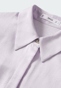 Chemise mauve clair avec col, présentant une texture lisse et un petit bouton au niveau du col, avec une étiquette indiquant la marque et la taille.