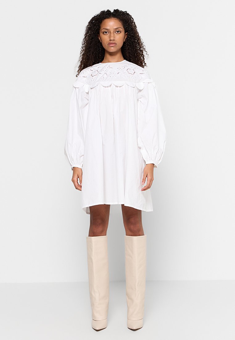 Vestido blanco oversize con un yugo bordado en forma de escollera y mangas amplias; combinado con botas de cuero crema hasta la rodilla.