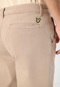 Beige bomullsbukser med smal passform, med en baklomme og en gul og svart brodert logo på høyre side.