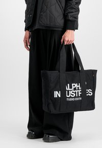 Czarna torba na ramię z podwójnymi uchwytami, z wyraźnym białym napisem "ALPHA INDUSTRIES STUDIO EDITION" z przodu. Płaski, prostokątny kształt.
