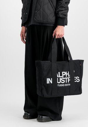 Črna platnena torba z dvojnih ročaji, na sprednji strani z izrazitim belim besedilom "ALPHA INDUSTRIES STUDIO EDITION". Ravna, pravokotna oblika.