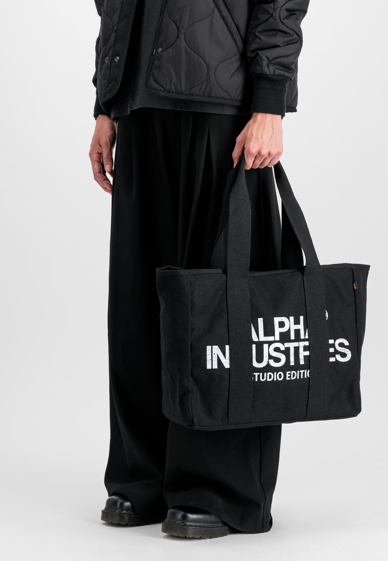 Czarna torba na ramię z podwójnymi uchwytami, z wyraźnym białym napisem "ALPHA INDUSTRIES STUDIO EDITION" z przodu. Płaski, prostokątny kształt.