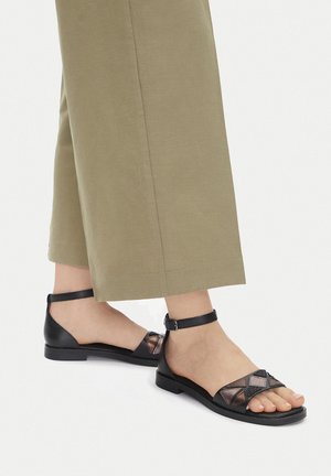 Sandalias planas negras con recortes geométricos, diseño de malla texturizada, correa en el tobillo y tacón bajo.