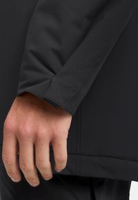 Gros plan sur une veste noire montrant une manche texturée avec un poignet ajusté. Le poignet comporte une petite boucle et des détails de couture soignés.