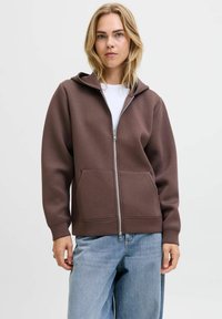 Felpa con zip marrone, dotata di cappuccio regolabile con cordoncino, tasche frontali e polsini a coste, abbinata a jeans blu scoloriti. Tessuto morbido, vestibilità casual.