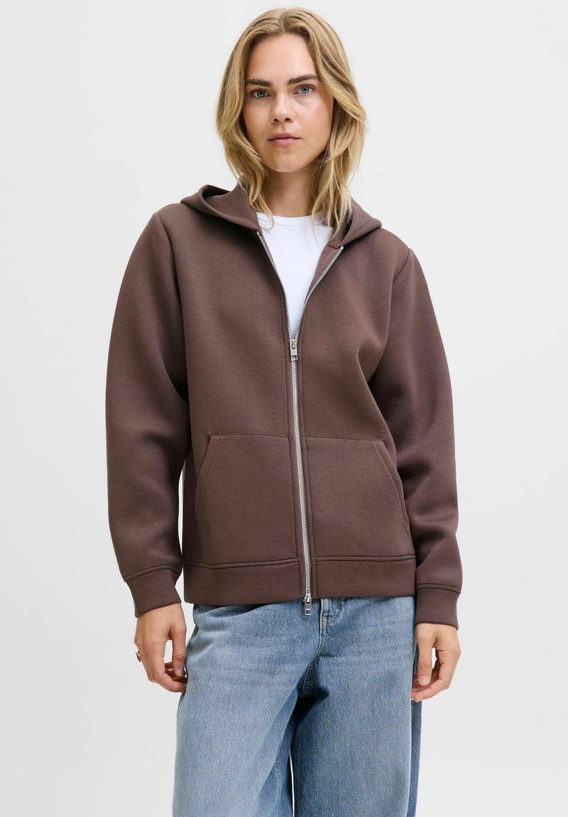 Felpa con zip marrone, dotata di cappuccio regolabile con cordoncino, tasche frontali e polsini a coste, abbinata a jeans blu scoloriti. Tessuto morbido, vestibilità casual.